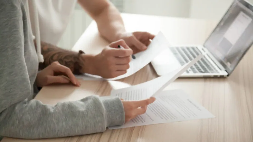 Duas pessoas sentadas numa mesa de madeira, na mesa estão alguns papéis um computador portátil. A imagem visa representar a análise de uma proposta de crédito