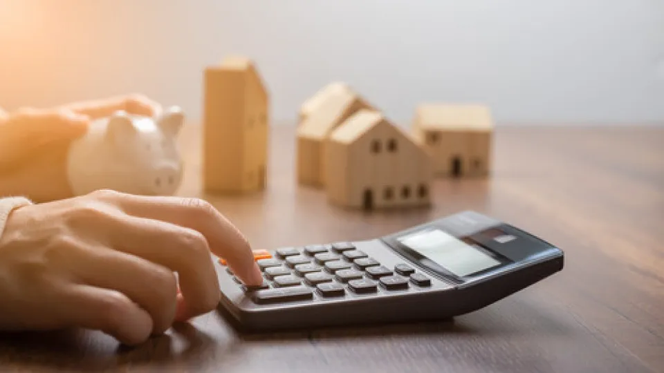 Pessoa a usar uma calculadora com modelos de casas e um mealheiro ao fundo, representando o cálculo da capacidade financeira para crédito habitação.