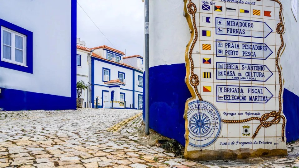 Rua típica da Ericeira com casas brancas e azuis e painel de azulejos com indicações turísticas.