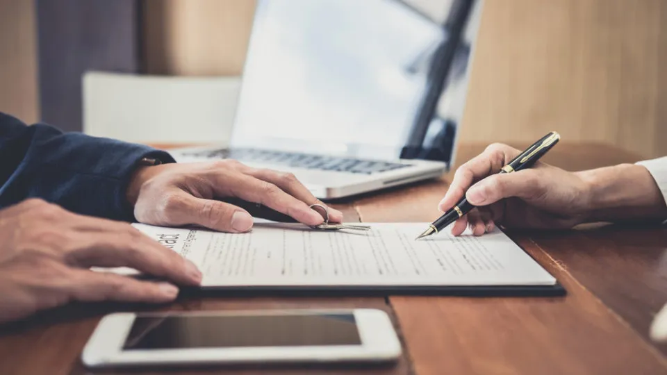 Duas pessoas a assinar um contrato sobre uma mesa, com portátil e telemóvel ao lado.