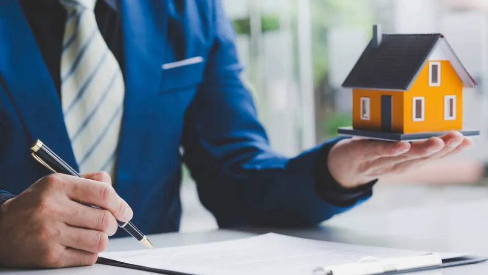 Profissional a analisar documentos com uma maquete de casa, representando a venda de um imóvel