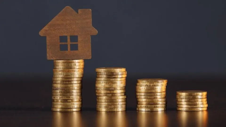 Casa simbólica sobre pilhas de moedas, representando os custos elevados e o financiamento na construção de habitação.