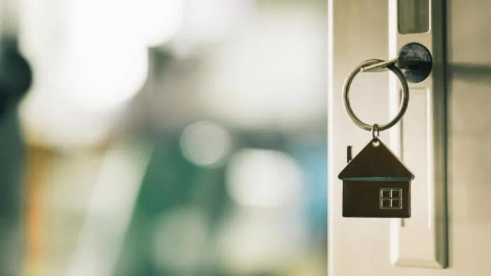 Chave com porta-chaves em forma de casa inserida numa fechadura, com fundo desfocado, simbolizando aquisição de imóvel ou mudança de habitação.