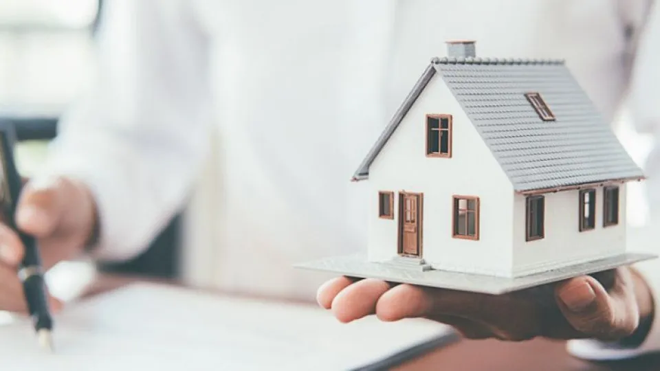 Pessoa segura uma maquete de casa numa mão e assina documentos com a outra, representando a reserva ou compra de um imóvel.