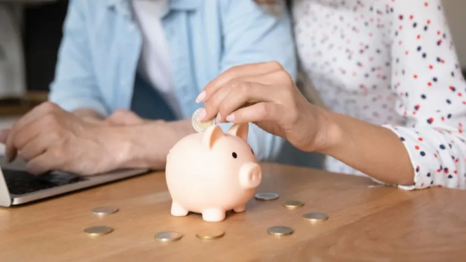 Pessoa a colocar uma moeda num mealheiro em frente a um portátil, simbolizando poupança e gestão financeira inteligente.