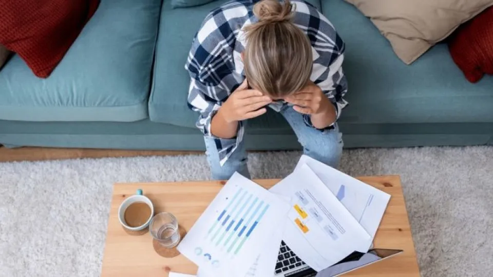 Mulher sentada em sofá com mãos na cabeça inclinada sobre mesa com papéis e portátil