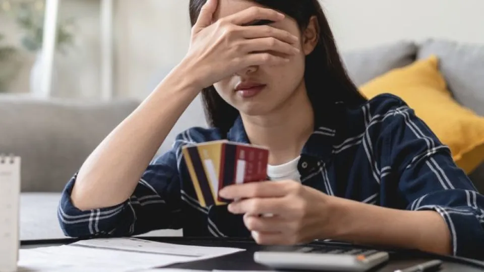 Mulher sentada no chão debruçada sobre mesa com mão na nos olhos e mão com cartões de crédito para simbolizar dívida às Finanças