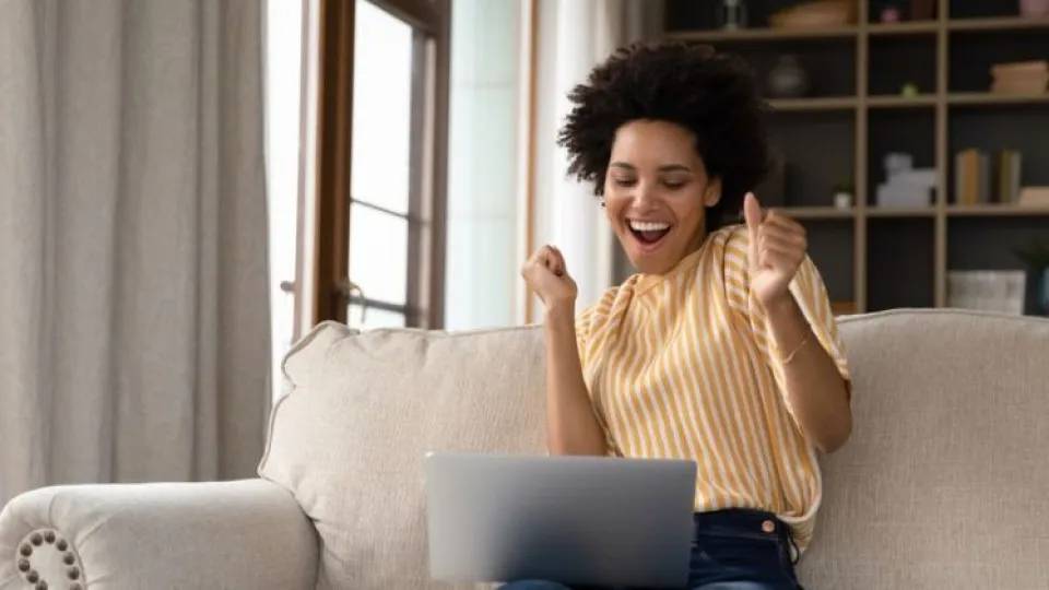 Mulher sentada em sofá com portátil ao colo e feliz a simbolizar o seu signo a ter sorte ao dinheiro