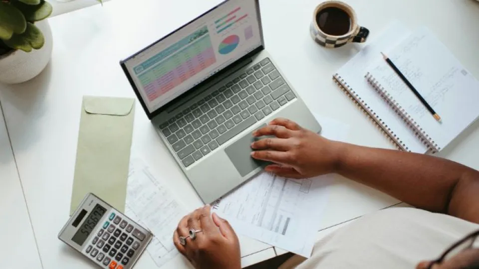 Mulher em secretária ao computador com estatísticas na tela e em folhas mais calculadora para simbolizar dados do salário médio baixo de Portugal