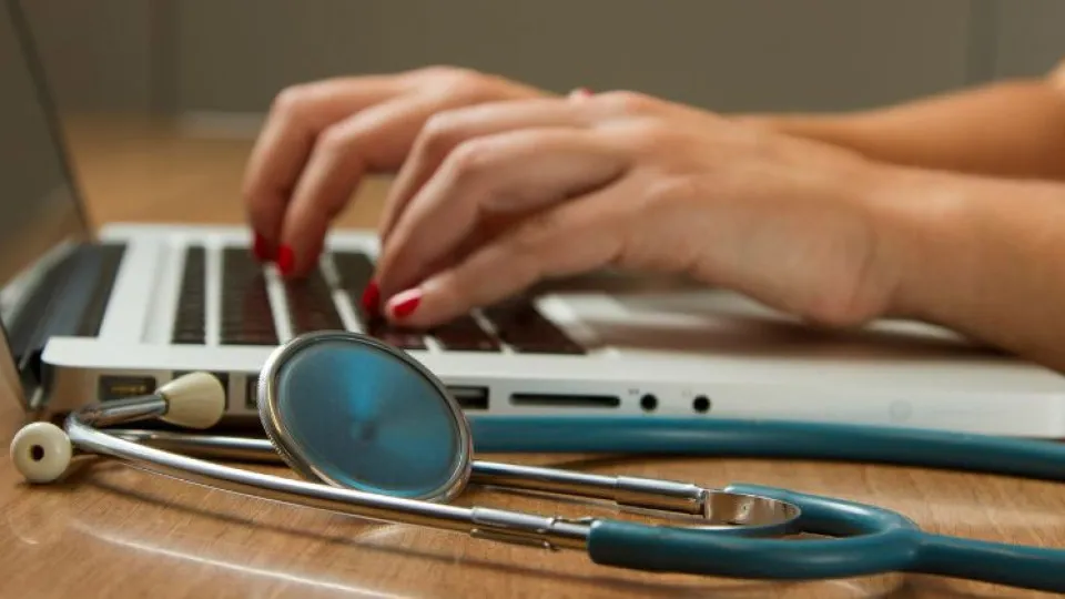 Mãos de senhora a escrever em portátil com estetoscópio ao lado