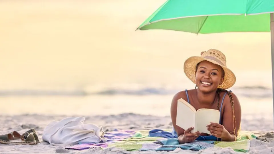 Senhora deitada em toalha na praia a ler livro sob chapéu de sol simbolizando férias poupadas em destinos baratos