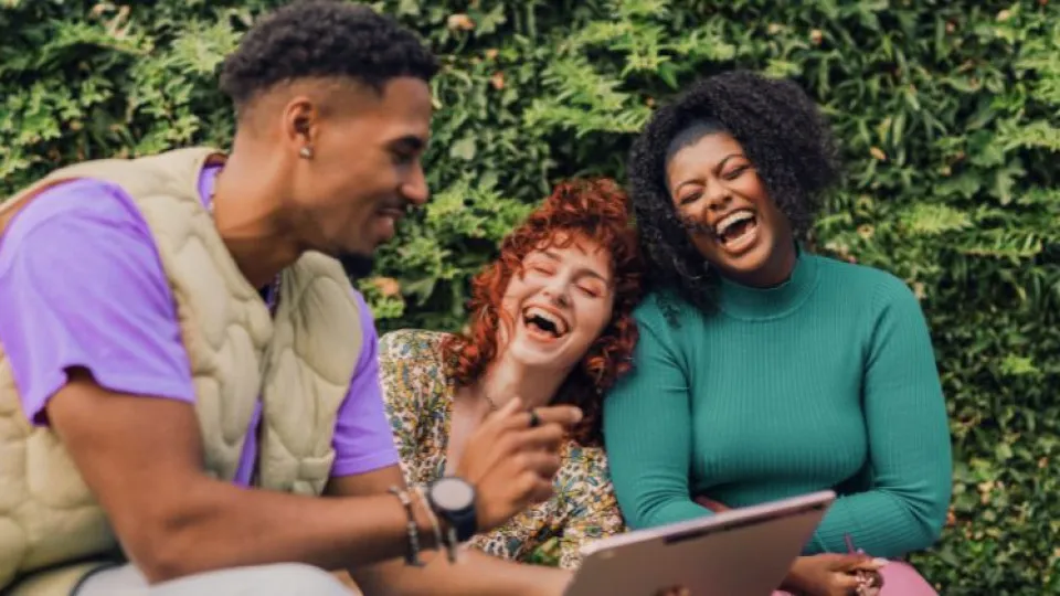 Três jovens adultos a rir enquanto olham para um tablet, sentados ao ar livre junto a uma sebe verde.
