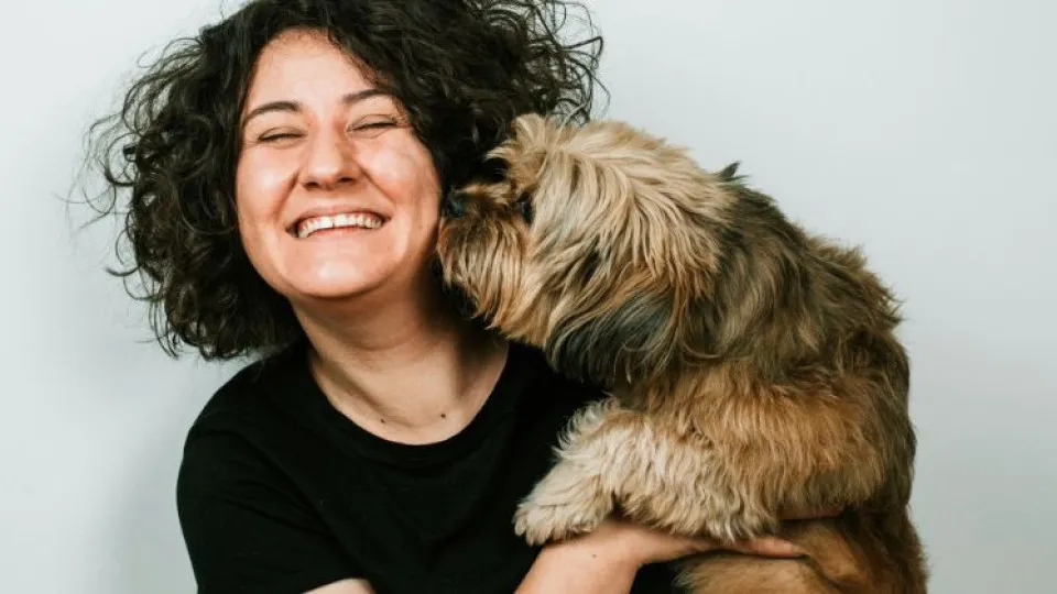 Mulher sorridente a segurar um cão de pequeno porte que lhe dá uma lambidela na cara, num momento de afeto e alegria.