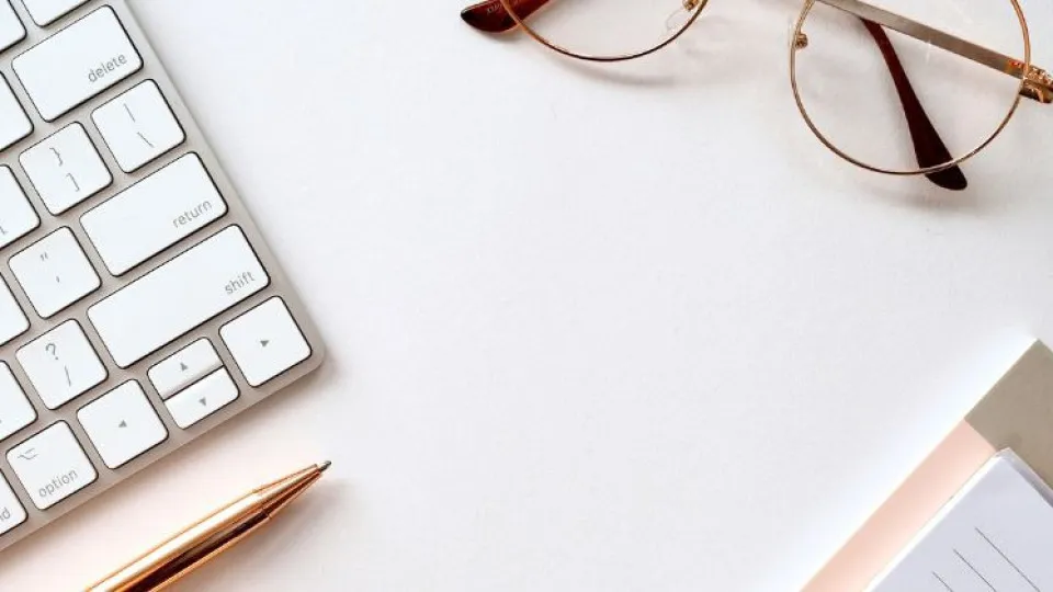 Secretária com teclado branco, caneta dourada, óculos e bloco de notas - ambiente de trabalho minimalista e organizado.