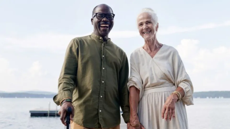 Casal de idosos sorridente a caminhar junto a um lago, de mãos dadas e com ar descontraído.