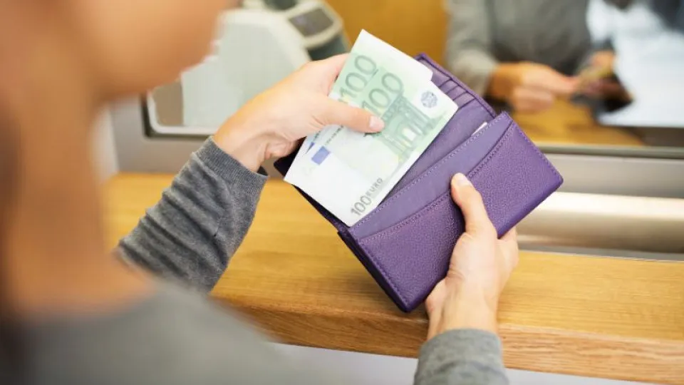 Pessoa a guardar notas de 100 euros numa carteira roxa ao balcão de um balcão de atendimento.