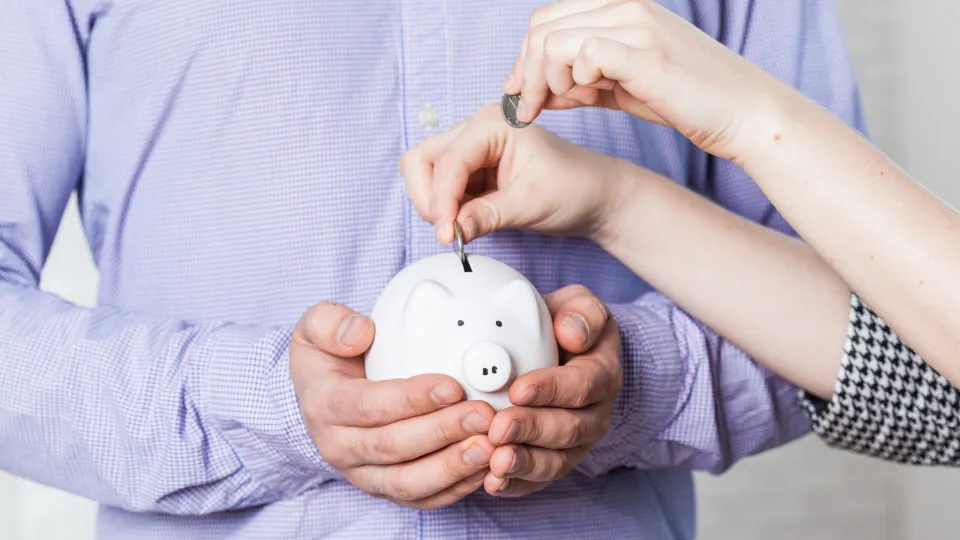 Duas pessoas a colocar uma moeda num mealheiro, simbolizando hábitos de poupança no dia a dia