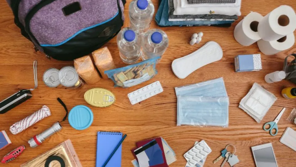 Conteúdo de um kit de emergência de 72 horas disposto no chão, incluindo água, alimentos enlatados, lanternas, medicamentos, documentos, máscara cirúrgica, artigos de higiene, papel higiénico, bloco de notas e objetos pessoais