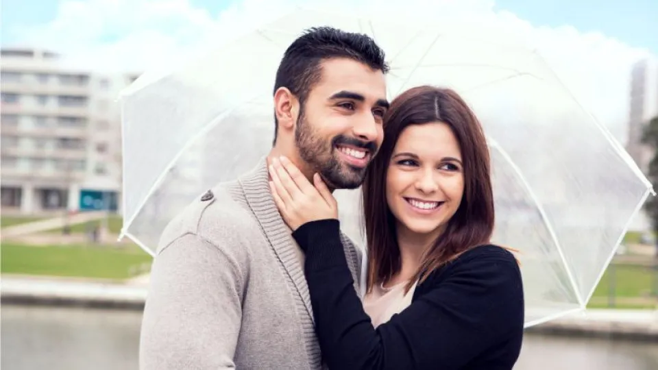 Casal sorridente abraçado debaixo de um guarda-chuva transparente num dia nublado, junto a um canal urbano.