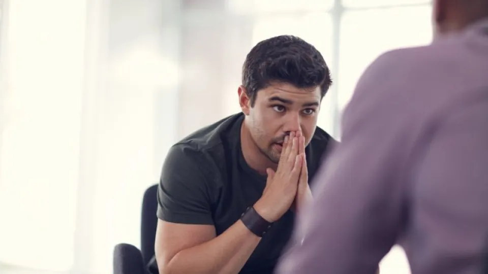 Homem jovem com expressão preocupada, sentado numa cadeira com as mãos junto ao rosto, aparentemente a conversar com outra pessoa num ambiente interior iluminado.