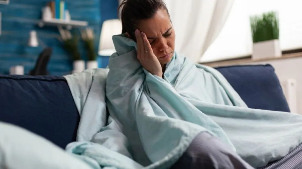 Mulher com doença em sofá enrolada em manta com mão na cabeça