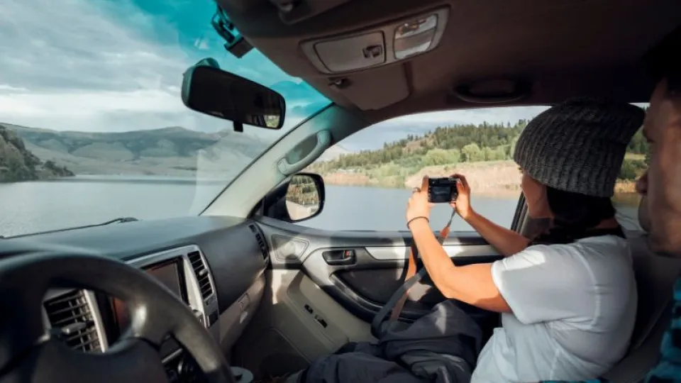 Casal dentro de um carro estacionado junto a um lago, com a mulher a tirar uma fotografia da paisagem através da janela, durante uma viagem de carro por zona montanhosa.