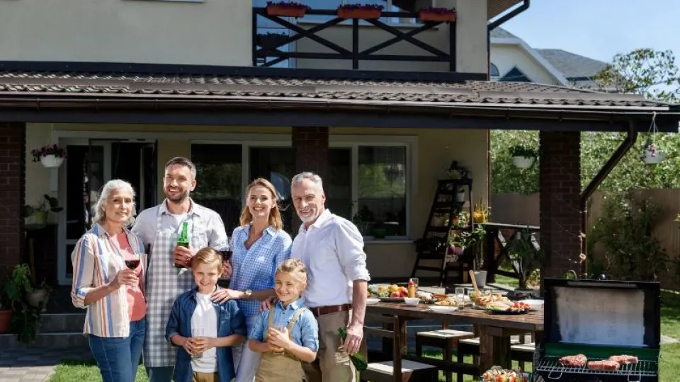 Família multigeracional reunida num churrasco no jardim de uma casa, sorrindo para a câmara, com mesa posta e grelhador ao fundo. Ambiente descontraído e ensolarado de verão.