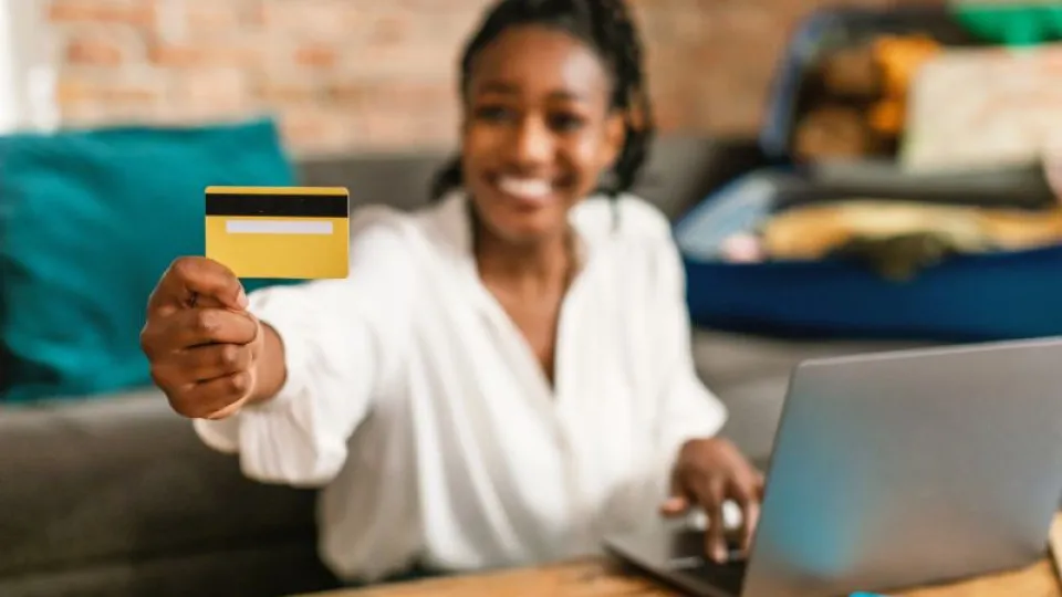 Mulher sorridente segura um cartão bancário amarelo enquanto usa um computador portátil.