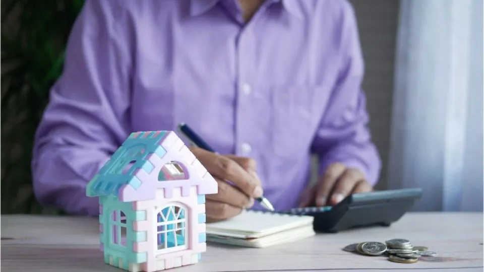 Homem de camisa roxa sentado à mesa com caneta na mão a escrever em caderno e com calculadora, maquete de casa mini e moedas na mesa