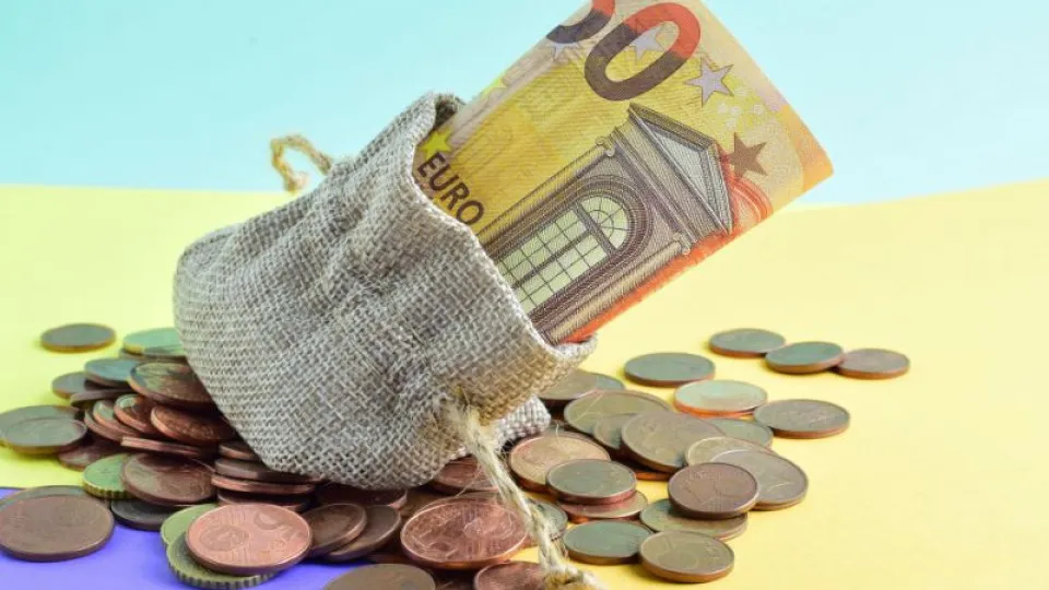 Saco de tecido rústico com uma nota de 50 euros parcialmente visível e várias moedas de euro espalhadas sobre uma superfície colorida.