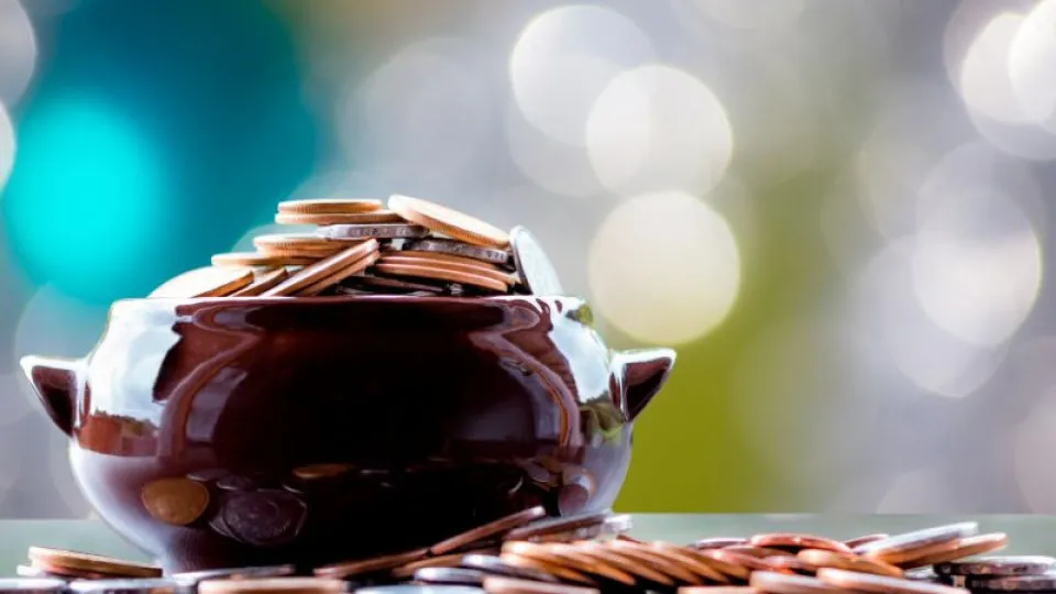 Cofre escuro cheio de moedas, com várias moedas espalhadas à sua volta sobre uma superfície, em frente a um fundo desfocado com luzes circulares suaves.