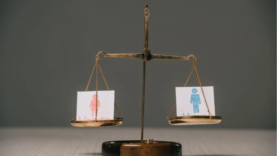 Balança de justiça com dois pratos: à esquerda, um cartão com a figura de uma mulher desenhada a vermelho; à direita, um cartão com a figura de um homem desenhada a azul. Ambos estão equilibrados, simbolizando igualdade de género.