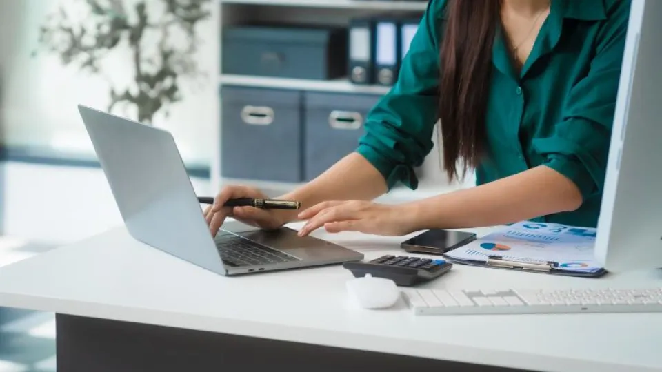 Mulher a trabalhar num computador portátil com documentos, gráficos e calculadora sobre a mesa, simbolizando gestão financeira ou análise de despesas.