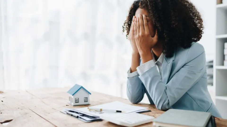 Mulher com ar preocupado a tapar o rosto com as mãos, sentada à secretária com documentos e uma miniatura de casa, representando dificuldades financeiras ou stress com crédito à habitação.