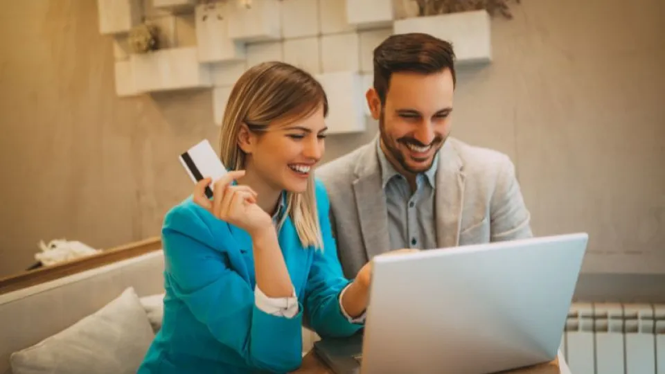 Casal jovem e sorridente usa computador portátil, com a mulher a segurar um cartão bancário, sugerindo uma compra ou operação financeira online num ambiente confortável e moderno.