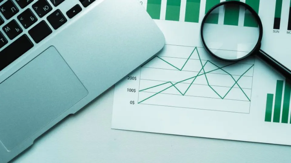 Portátil prateado parcialmente visível sobre uma mesa branca, ao lado de folhas com gráficos de barras e linhas em tons de verde. Uma lupa preta está posicionada sobre os gráficos, sugerindo análise de dados financeiros.