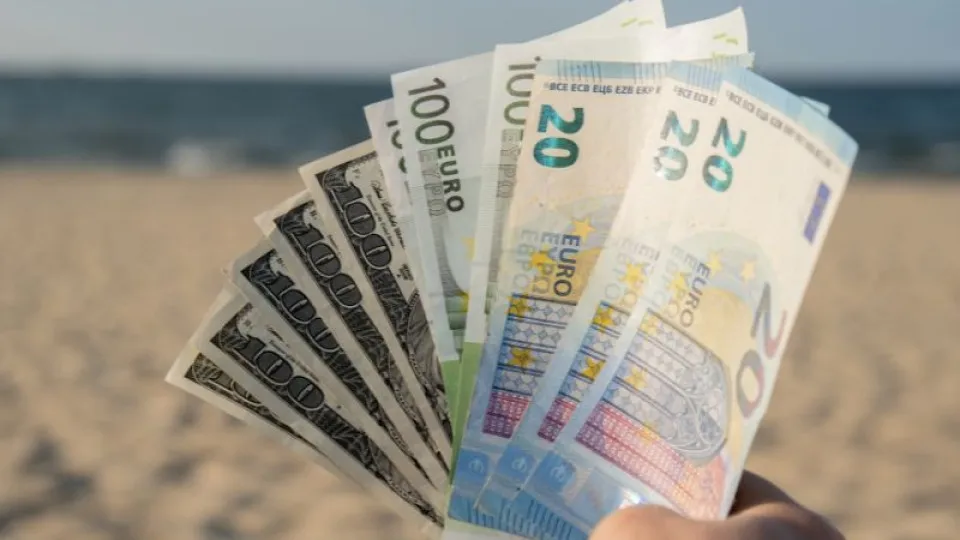 Mão a segurar várias notas de euro e dólares americanos em leque, com fundo de praia e mar desfocado ao longe, simbolizando férias e gestão de dinheiro.