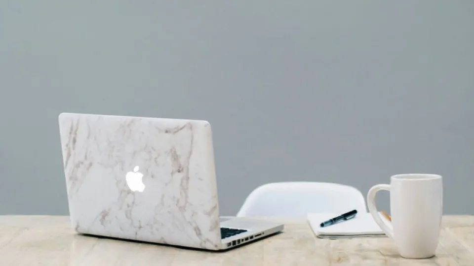 Mesa de madeira clara com um laptop com capa de mármore, um caderno com uma caneta preta em cima e uma caneca branca, em frente a uma cadeira branca, com fundo cinza liso.