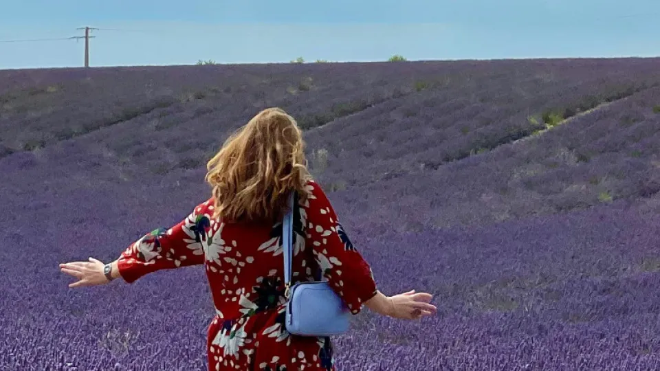 Mulher de costas num campo de lavanda, com braços abertos e vestido florido, a aproveitar um momento de tranquilidade ao ar livre.