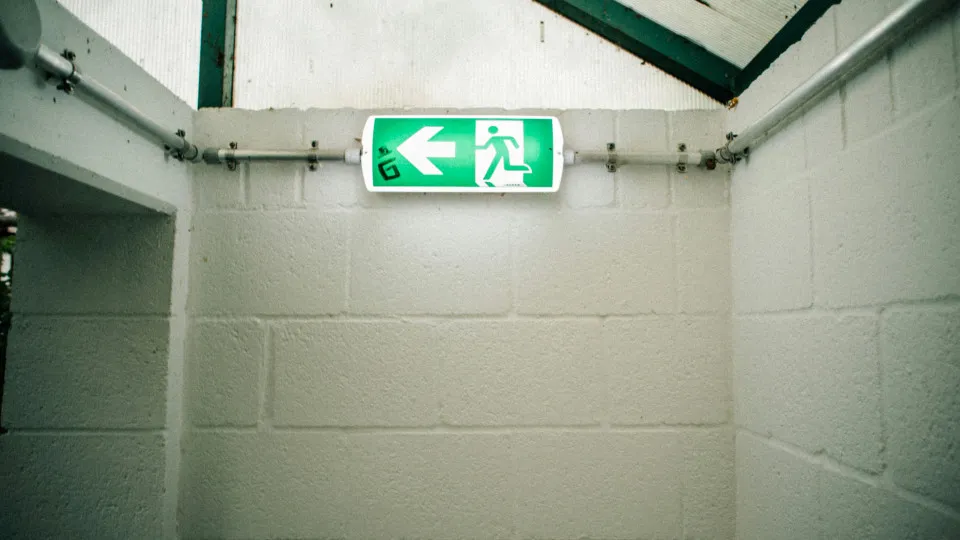 Sinal verde de saída de emergência iluminado, apontando para a esquerda, numa escadaria interior com paredes brancas de blocos de cimento.