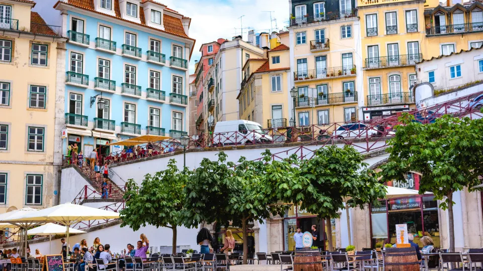 Vista de uma esplanada com árvores e barris de vinho na Praça do Município, em Lisboa, rodeada por prédios típicos e coloridos com escadaria ao fundo.