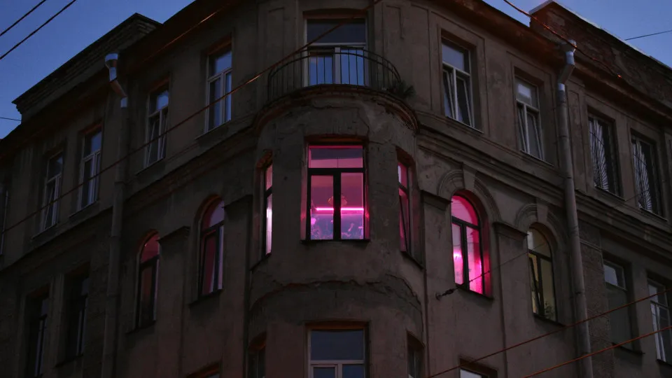 Fachada de prédio antigo ao anoitecer, com luzes cor-de-rosa acesas no interior de alguns apartamentos.