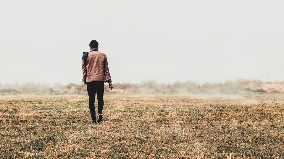 Homem de costas a caminhar num campo seco com uma criança ao colo, envoltos por neblina ao fundo. Cena transmite solidão, mudança ou reflexão.