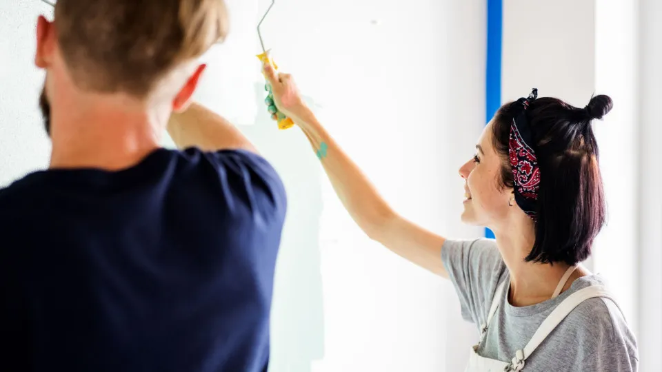 Casal jovem a pintar uma parede de branco e verde claro, juntos, durante obras de renovação numa casa.