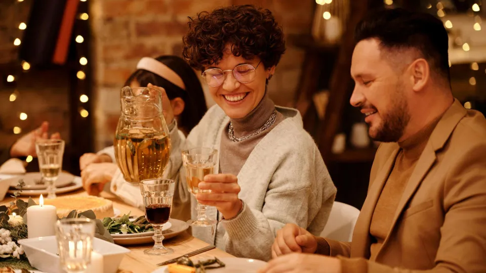 Grupo de pessoas a celebrar à mesa com bebidas e decoração natalícia, simbolizando a ceia de Natal e a importância de poupar nos preparativos.