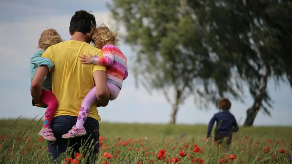 Pai a caminhar num campo com os filhos ao colo, simbolizando proteção familiar proporcionada pelo seguro de vida.