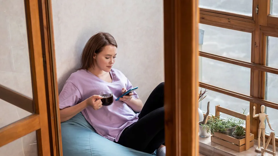 Mulher sentada junto à janela, com café na mão e telemóvel, a organizar as finanças e planear o início do ano.
